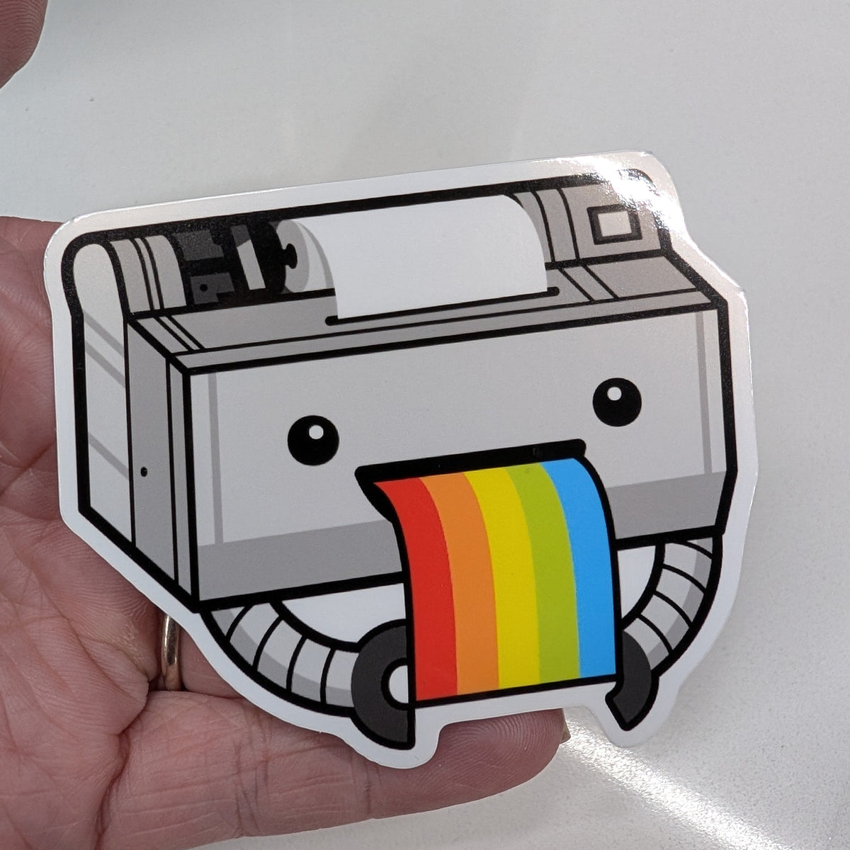 A die-cut sticker of a cute, cartoonish printer with a rainbow-colored paper strip coming out of its mouth, held in a person's hand against a white background.