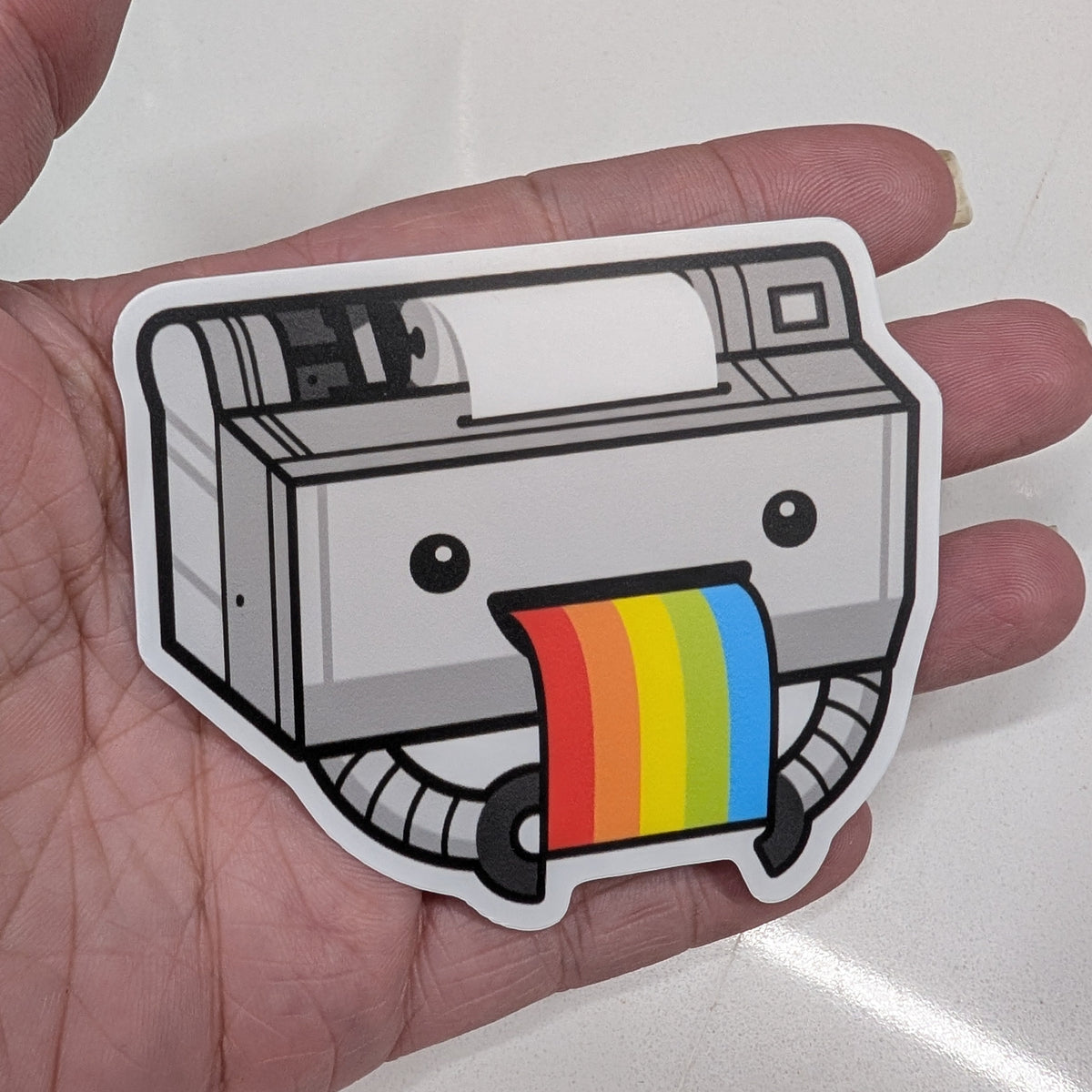 A die-cut sticker of a cute, cartoonish printer being held in a person's hand. The printer is gray and black with a colorful rainbow-striped paper output.